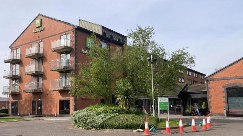 On the left is a four-storey red brick building with glass balconies. On the building is a green Holiday Inn logo and signage. In the middle is a landscaped lawn which is half surrounded by red and orange cones.