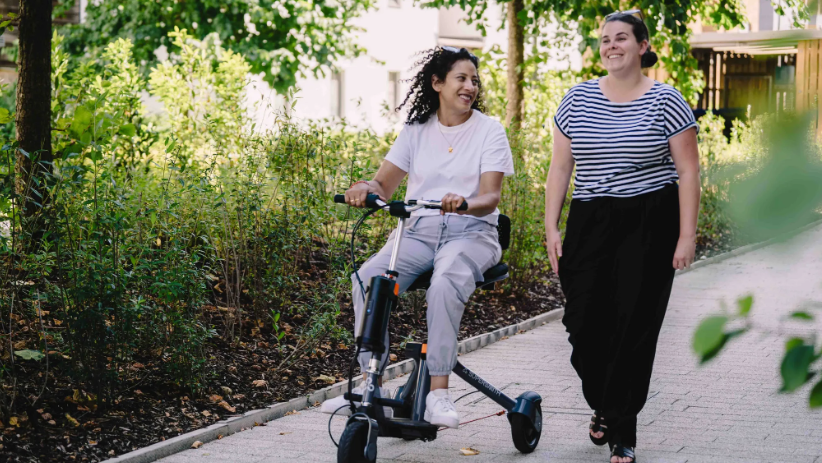 A woman riding a three wheeled scooter and talking with another woman who is walking by her side