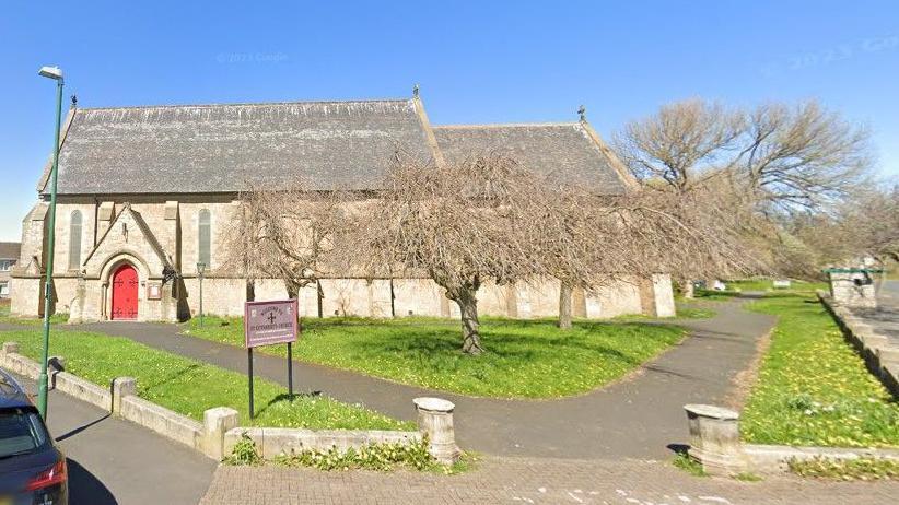 St Cuthbert's Church in Hebburn. It is an old-looking stone-coloured church with large trees outside. The trees have no leaves. The church has a red door. The grounds have grass. It has long glass windows.