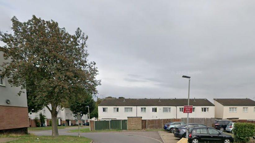 A complex of two-storey flats, painted white with grey roofs. There is a fence in front and a car park in the foreground. There is a tree to the left.