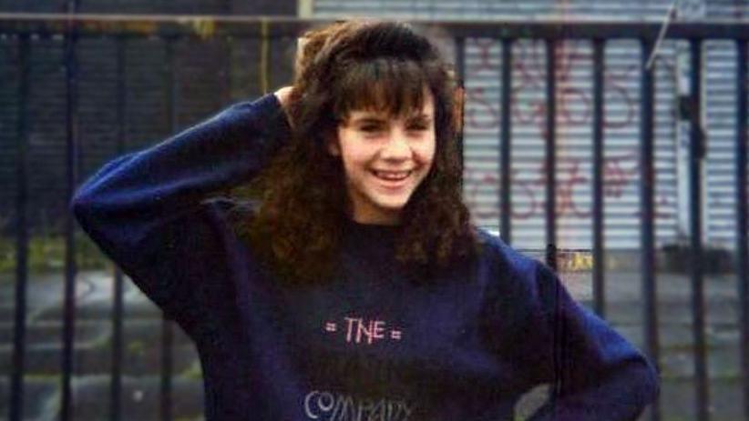 A photo of Caroline Glachan. She is a teenager posing outside in front of an iron railing. Behind the rail is a set of iron shutters covered in graffiti. Caroline is wearing a blue sweater and smiling at the camera. She has one hand behind her head. 