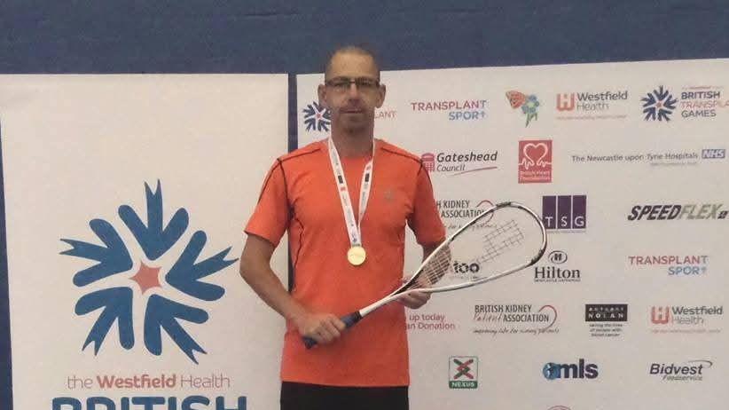 A man in an orange T shirt stands against a media board holding a squash racket. He has a gold medal around his neck. He is wearing rectangular glasses.