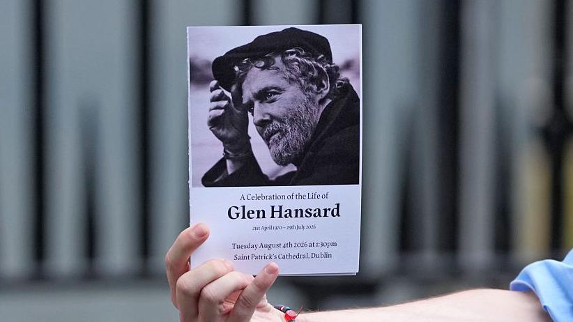 An arm, with the hand holding up a small booklet. The booklet has a black and white photo of a man with a beard on the front. Below the photograph are the words: 'A Celebration of the Life of Glen Hansard'. 