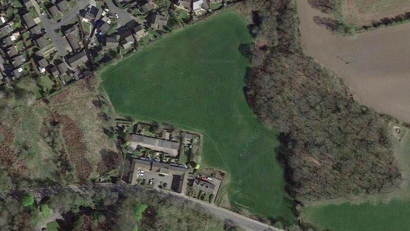 Aerial view, a field in the centre, woodland lining it to the right. Either side of that there are a few houses, they look like farm buildings, and other fields which appear brown/beige from above.