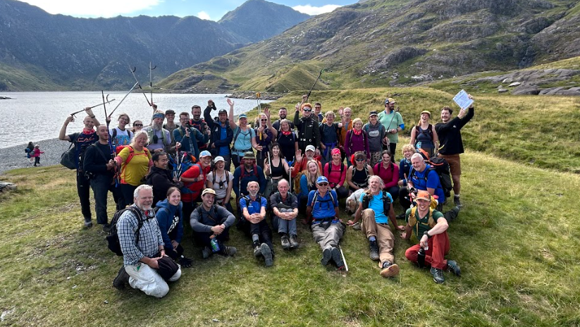 A large group of people in outdoor gear, holding up their hands and litter pickers and smiling at the camera. Behind them is a stunning mountain landscape and a body of water.