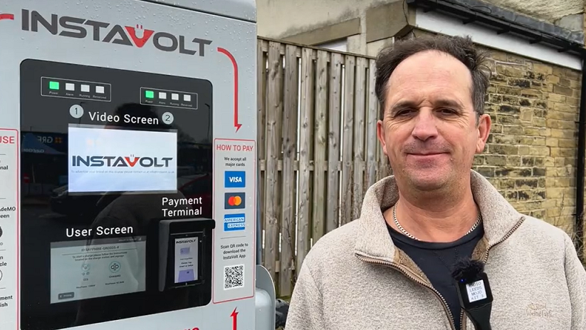 A brown-haired man man wearing a beige zip-up sweatshirt and blue t-shirt stands in front of an electric car charging point.