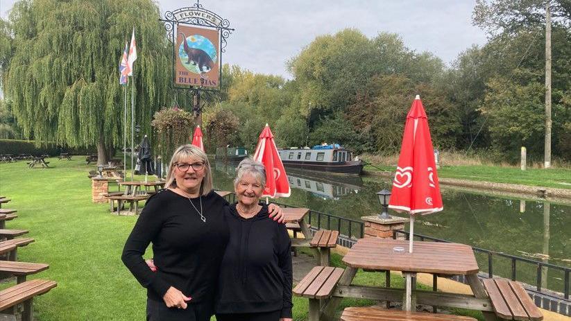 Jane Murphy and Margaret Stafford are hugging each other as they stand in the canal side beer garden of the pub. A sign with a picture of a dinosaur says 'The Blue Lias' and a canal boat can be seen moored on the canal behind them. Mother and daughter are smiling.