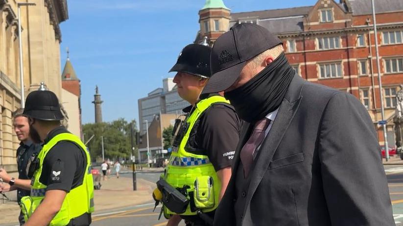 Robert Bush wears a dark grey suit, dark-grey cap and black scarf, which is wrapped around much of his face below his eyes. He is walking on a street accompanied by three police officers in high-visibility vests and black uniforms. Behind them is an old red-brick and stone building, with other buildings and a monument visible in the distance under a clear blue sky.