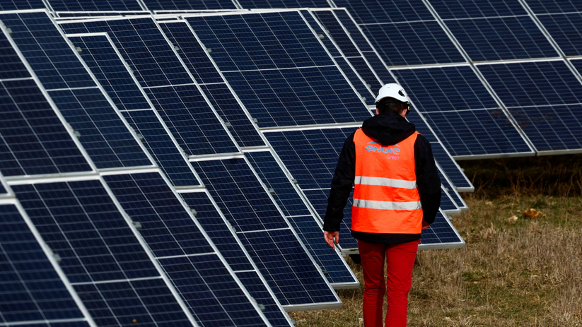 Man in white hard hat and orange hi vis vest walking away from the camera alongside several solar panels 