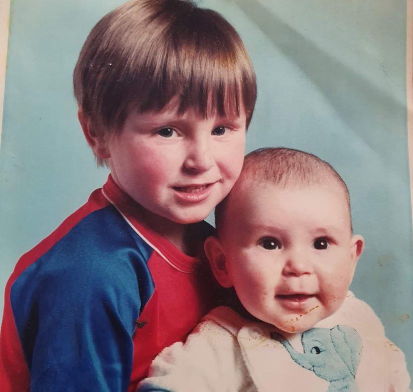 John Leech, wearing a blue and red t-shirt, holds his baby brother Danny. who is wearing a cream coloured bib featuring a picture of an elephant. 