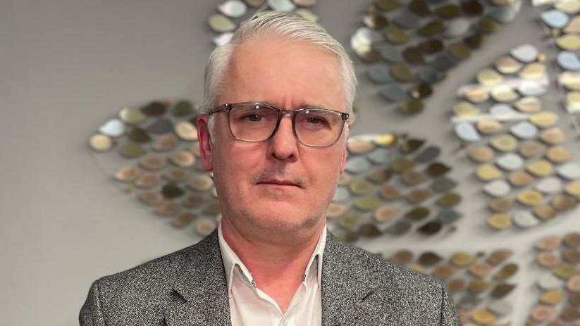 A man in his 50s looks with a serious expression into the camera. He wears a white shirt with the top button undone, grey and black blazer, and glasses. He has silver hair.