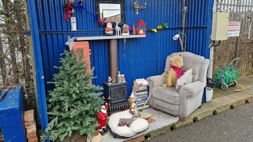 The inside of a blue, corregated metal alcover featuring a cozy-looking lounge. A stuffed dog sits by a log burner in an armchair.  A christmas tree stands next to the fire and there is a mirror on the wall.