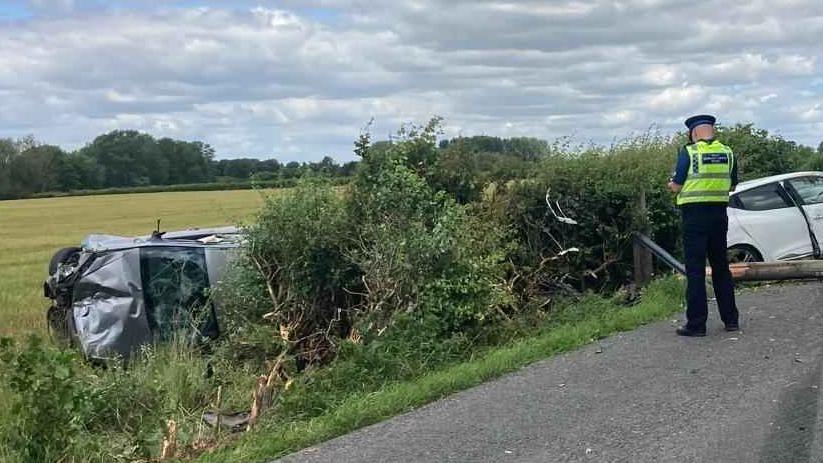 A crash site on a country road. A silver car has rolled over and is crumpled in a field, by a hedge. There is a police officer and a white damaged car on the right.