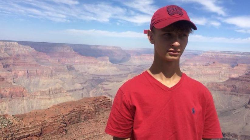Young man wearing a red T-shirt and a red cap standing in front of the Grand Canyon. He is looking off camera.