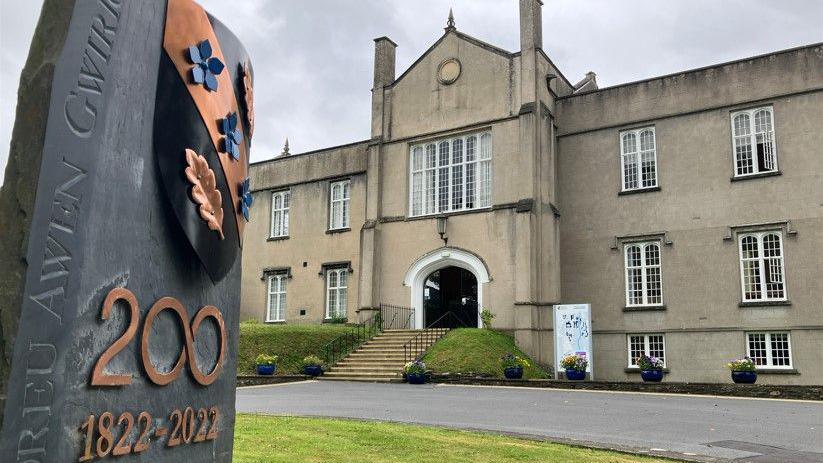 The historic stone Lampeter campus of the University of Wales Trinity St David building, with stone steps leading up to a large arched entrance. There are multiple mullioned windows. A slate sign in front of the building has copper coloured lettering which says '200' and '1822-2022'.