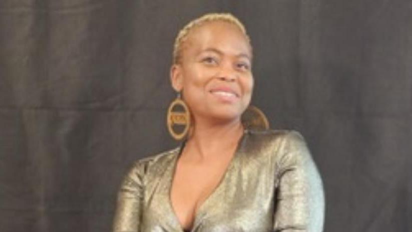 Shows a woman in a sparkly dress, cropped golden hair and large round golden earrings.
