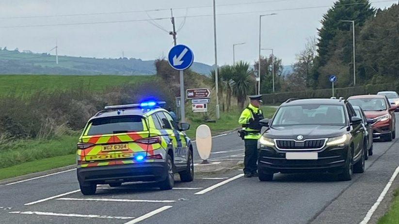 A police checkpoint on a road. There is a police car parked in the middle of two lanes. There is a line of traffic in the right hand lane and a police officer is taking to the person in the car at the front of the queue, a black Skoda. 