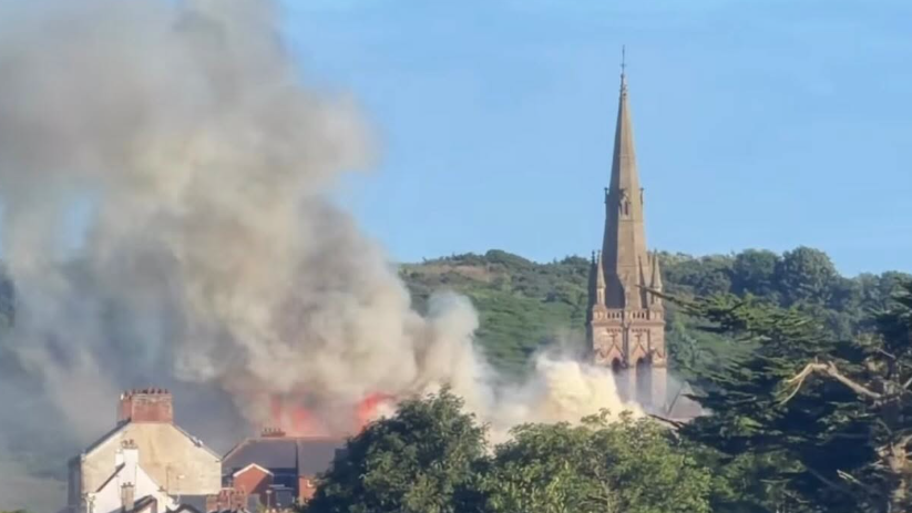 A horizon of the tops of buildings and green trees. A towering grey church spire sits above the rest of the buildings. Grey smoke is bellowing from the trees to the left of the church. 