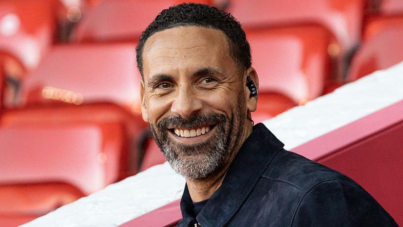 A man is looking at a camera and smiling. He has short, dark hair, a beard and is wearing a black jacket. Behind him there are rows of red seats at a football stadium.