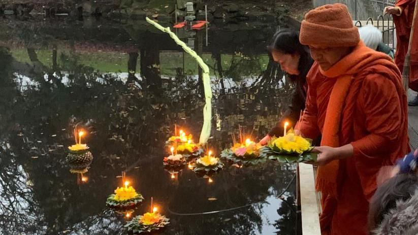 The Venerable wears orange drapes and an orange hat, holding a krathong with green banana leaves and orange flowers and a candle in the middle. He is releasing it into a large pond of water. 