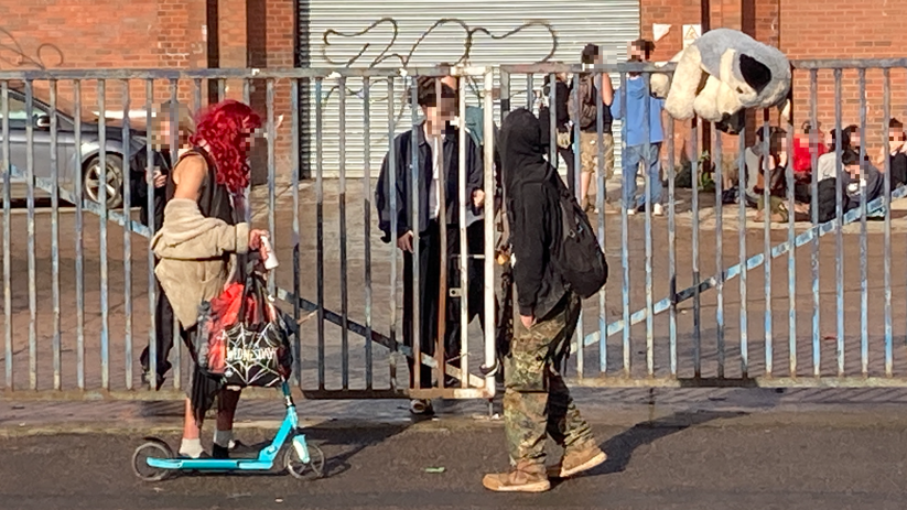 People around a warehouse, some dressed in fancy dress costumes, one person is riding a scooter. Their faces have been pixelated.