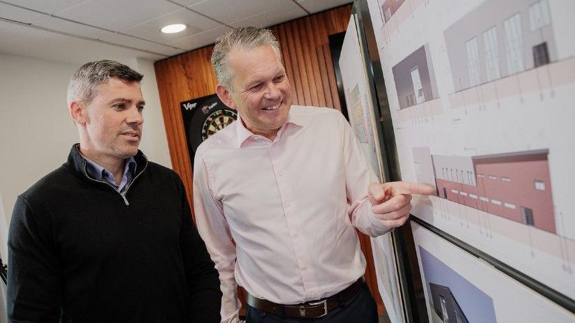 Two men, one wearing a black sweater and the other wearing a light pink shirt, look at construction plans which are pinned up on a wall. There is a dartboard and wood panelling behind them.