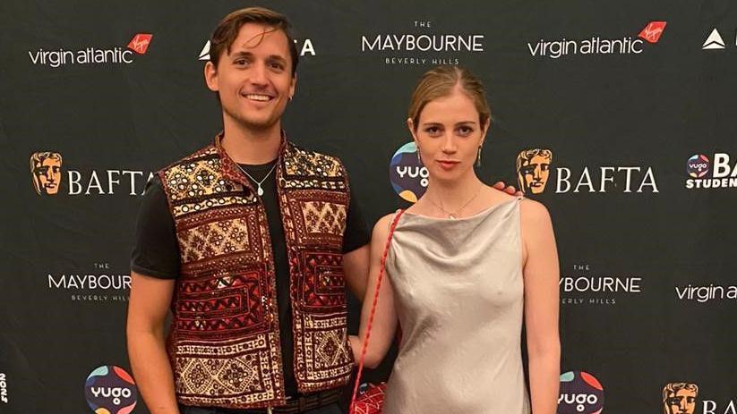 Nyal standing in front of a black background with BAFTA logos. He is wearing a colourful vest over a black tee shirt. Next to him is his partner Chloe wearing a grey dress