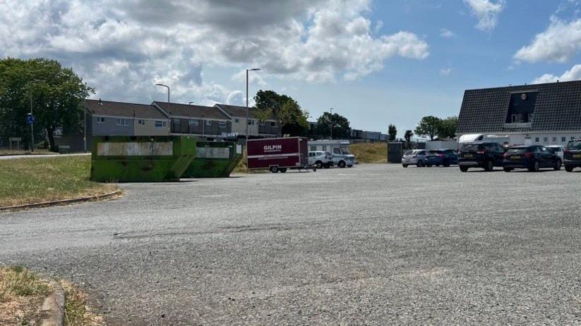 Windmill carpark with parked cars. The pub building in the distance with large green bottle bins to the left of the photograph