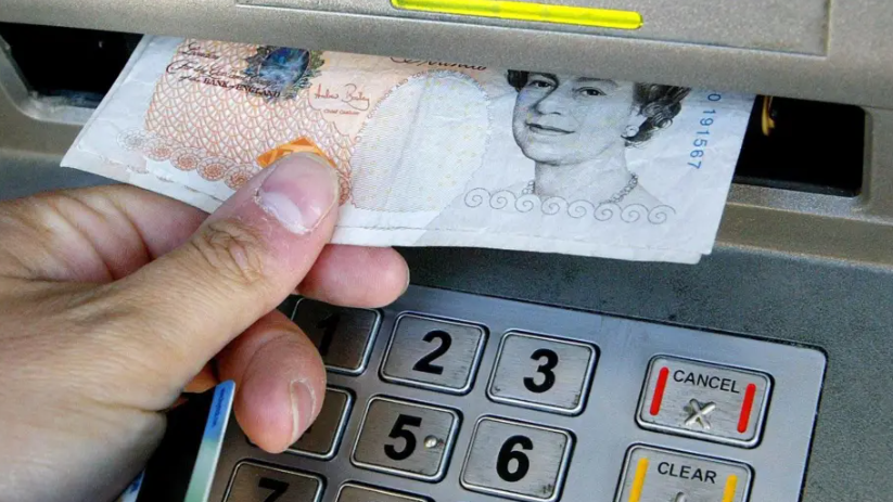 A stock image showing a hand withdrawing multiple £10 notes from an ATM. A silver keypad for PIN numbers can also be seen