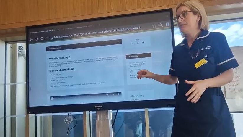 A MUMS Group midwife wearing a blue uniform standing in front of a large television screen