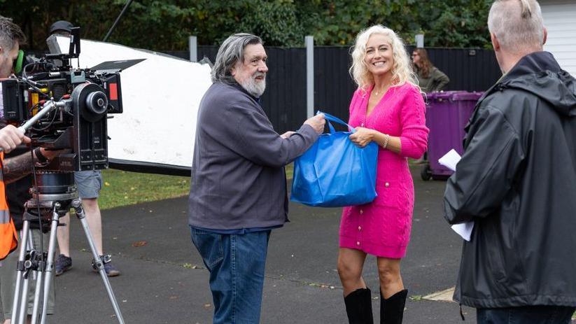 Ricky Tomlinson is pictured on set with Suzanne Collins on the residential street known as Brookside Close. He is wearing jeans and a grey cardigan and she is wearing a pink dress.