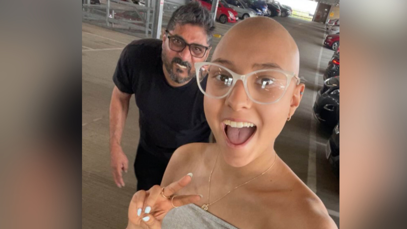 Asha with her father, Jayesh. They are walking in a multi-storey car park. Asha is wearing glasses and has a shaved head following her treatment. She has a shoulder-less silver dress on and has her mouth open while doing a peace sign with her fingers. Jayesh is crouched in the background smiling while wearing a black T-shirt and glasses.