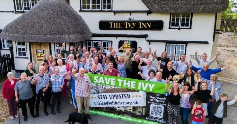 About 30 supporters - standing outside the Ivy Inn, which is a traditional old pub with whitewashed walls and thatched roof - all with their thumbs up in the air and smiling. They are holding a large banner saying 'Save the Ivy'.