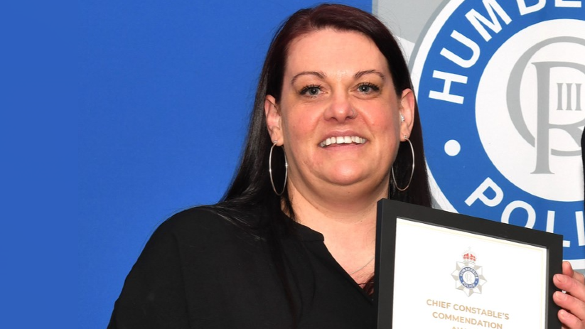 Jodie Offen, a woman with long dark hair, wearing a black blouse and silver hoop earrings, holding a black-framed award reading 'Chief Constable's Commendation'. It is a head and shoulders shot of Ms Offen looking and smiling into the camera. She is stood in front of a blue wall with a cut-off Humberside Police sign in the back left of the photo. 