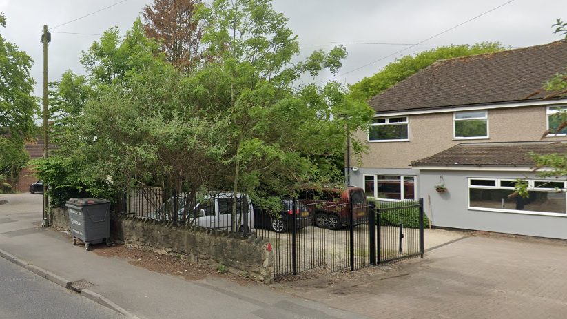 A suburban care home called Bobbins surrounded with black railings and cars parked on the drive. There are trees in the foreground and a stretch of pavement. 