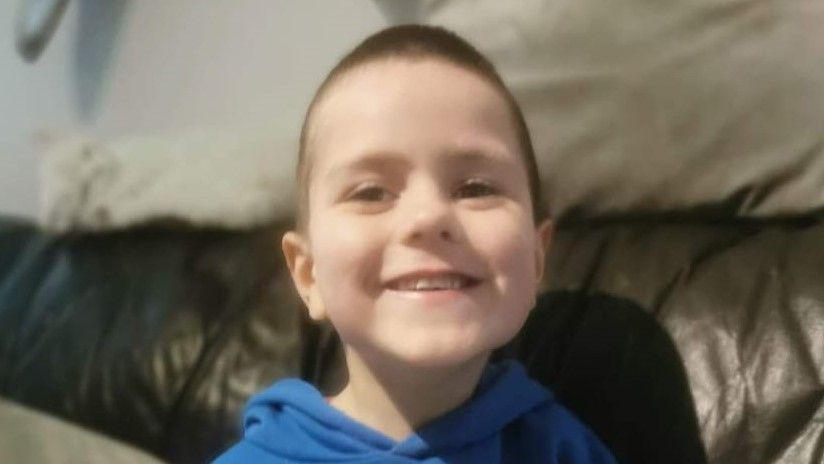 A young boy smiling at the camera. He is wearing a blue hoodie and sitting on a leather sofa.