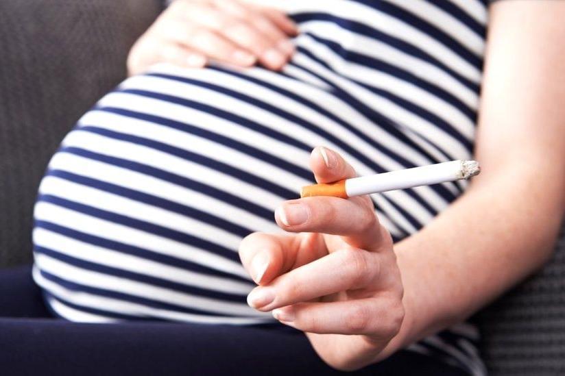 Pregnant woman in blue and white striped top with right hand resting on baby bump and left hand holding a lit cigarette.