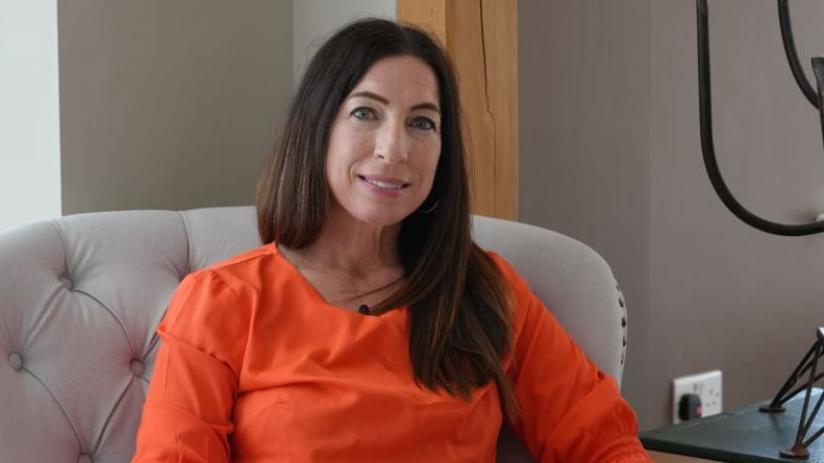 Dr Beth Mosley sits in a chair and looks towards the camera. She has long dark hair and wears an orange top.