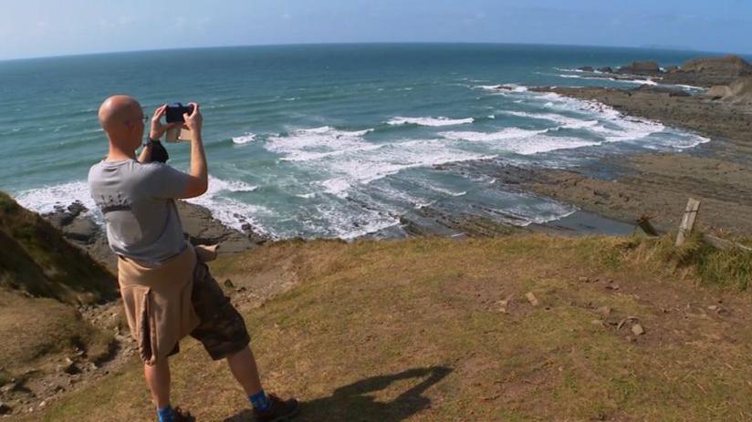 A person is pictured taking a picture of the coast line. He is facing away from the camera and is wearing a grey shirt with black shorts. In front of him is the coast. It is a blue coloured water and rocks can be seen breaking through the water.
