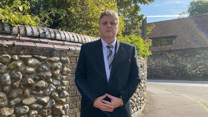 Man dressed in a suit standing in front of a flint wall his hands clasped in front of him