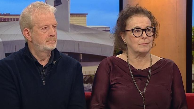 Charles and Liz Ritchie sit on the sofa on BBC Breakfast. Charles wears a blue jumper and Liz a red top and glasses. 