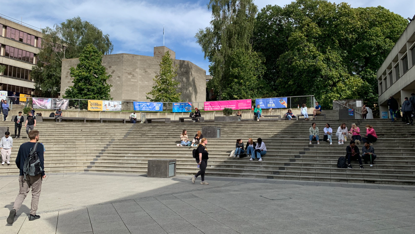 Outside the university buildings, with a flight of curved concrete steps and students sitting on them in groups