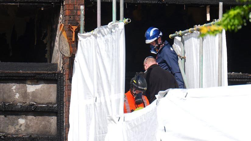 Three people, two wearing breathing apparatus are seen behind white sheeting during what appears to be a forensic investigation of the scene. The blackened building is seen to the left of them.