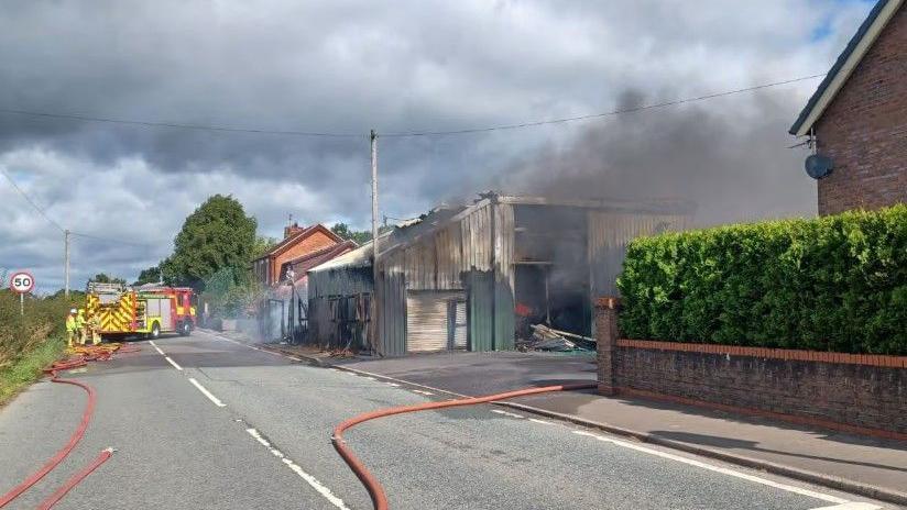 A green wooden building on fire, with black smoke rising from the building, as the green paint peels away. Orange firefighter hoses run across the street, with a fire engine in the background