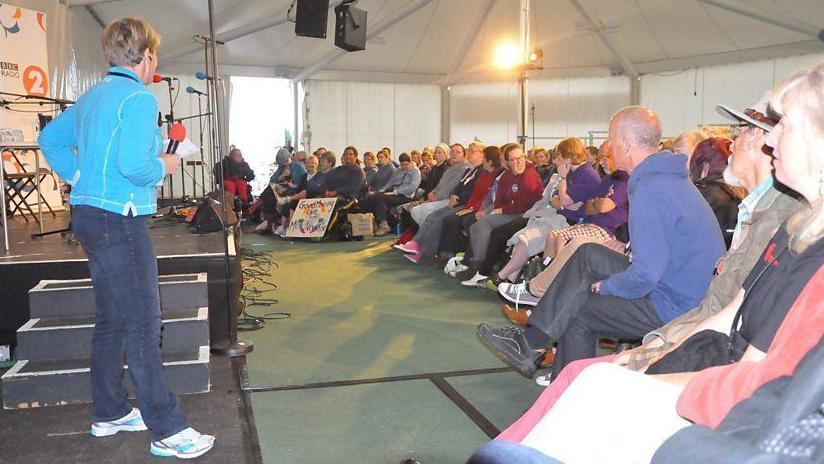 Claire Balding with medium-length blonde hair wearing a light blue top and dark blue trousers, standing beside the steps to a stage inside a marquee. She is holding a microphone with a red windshield. People are sitting in chairs facing her. There are two speakers suspended from the ceiling.