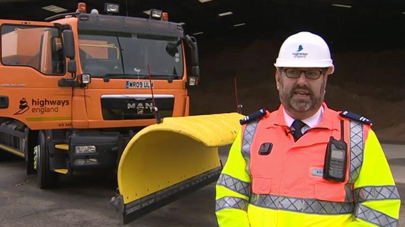 Mark Munnoch with dark hair, a beard and glasses wearing a white helmet and yellow and orange hi-vis jacket. He has a radio attached to his jacket. There is an orange Highways England lorry behind him that has a yellow snow plough on the front.