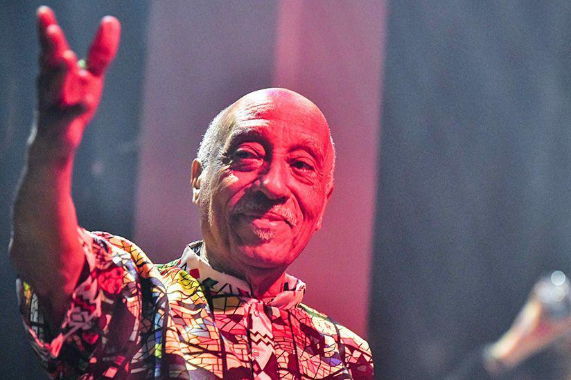 Mulatu Asatke wearing a patterned shirt smiles and waves to the audience.