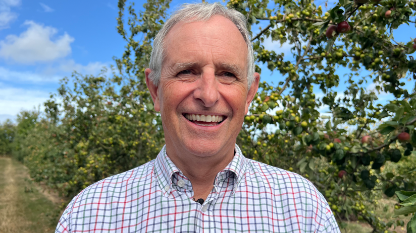Martin Thatcher is smiling at the camera while stood in one of the apple orchards at the farm's large site. He is wearing a white checked shirt with red and blue lines.