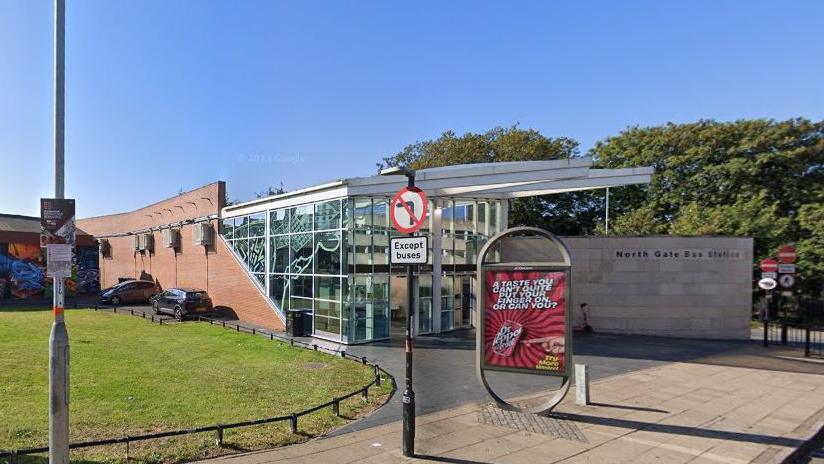 Glass-fronted building with stone wall adjacent bearing the legend "North Gate Bus Station". The rear of the building is faced in brick. There is a free standing advertising display stand on the pavement in the foreground promoting a soft drink, and a sign indicating no left turn except for buses. A grass plot is visible to the left.