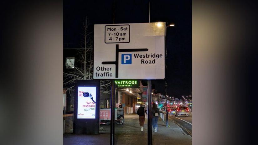 A road sign pictured at night. A left turn is labelled "other traffic", a right turn is labelled "Parking Westridge Road" and straight ahead is labelled with traffic restrictions indicating cars may not drive in that direction at certain times. 